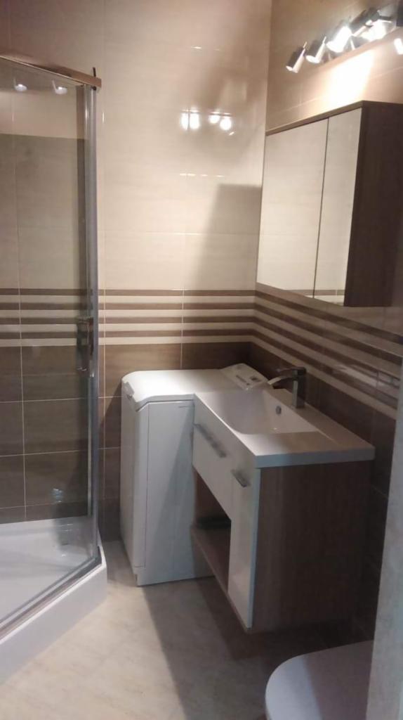 Nowoczesna łazienka z kabiną prysznicową, pralką pod blatem umywalki i szafką z lustrem, wykończona beżowo-brązowymi płytkami.