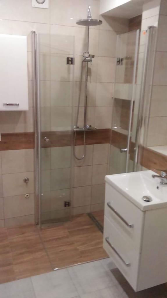 Nowoczesna kabina prysznicowa ze szklanymi drzwiami, deszczownicą i baterią termostatyczną, umywalka z szafką w białym kolorze, płytki w odcieniach beżu i brązu, widoczne elementy instalacji.