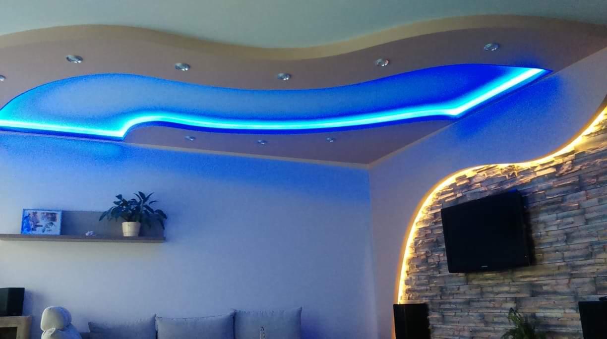 Sufit podwieszany z falistym kształtem i niebieskim oświetleniem LED, kamienna ściana z podświetleniem i telewizorem w salonie.