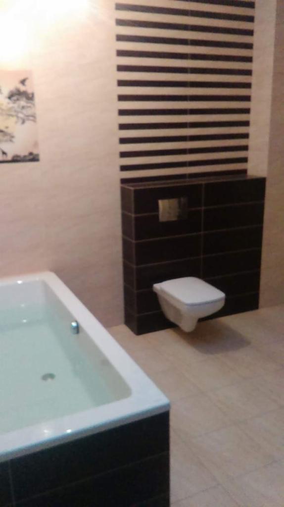 Nowoczesna łazienka z podwieszaną toaletą obudowaną ciemnymi płytkami, białą wanną wypełnioną wodą i dekoracyjnym panelem ściennym z poziomymi pasami.