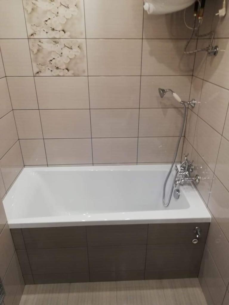 Biała wanna obudowana panelami z płytek imitujących drewno, kremowe płytki na ścianach z subtelnym motywem kwiatowym, chromowana armatura łazienkowa.