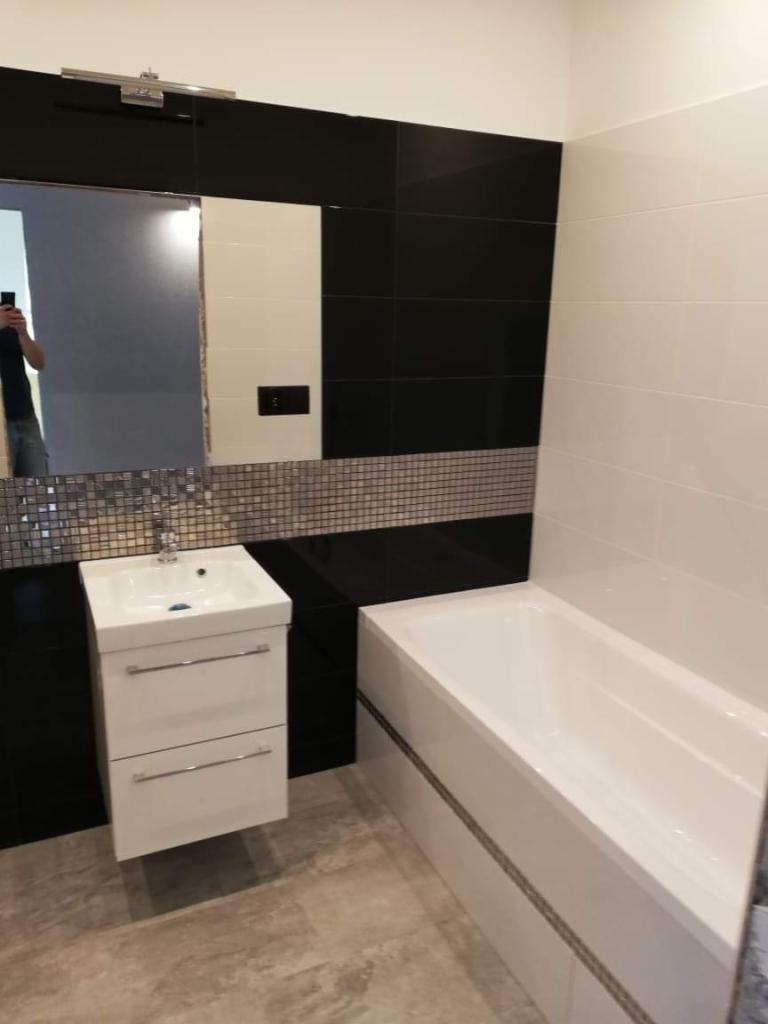 Nowoczesna łazienka z białą armaturą, czarnymi i białymi płytkami oraz mozaikowym akcentem, widok z perspektywy osoby robiącej zdjęcie w lustrze.