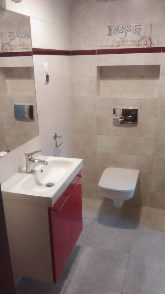 Wykończona łazienka z umywalką na szafce w kolorze czerwonym, toaleta podwieszana, beżowe płytki z dekoracyjnym pasem z motywem cyfr, lustro i akcesoria łazienkowe.