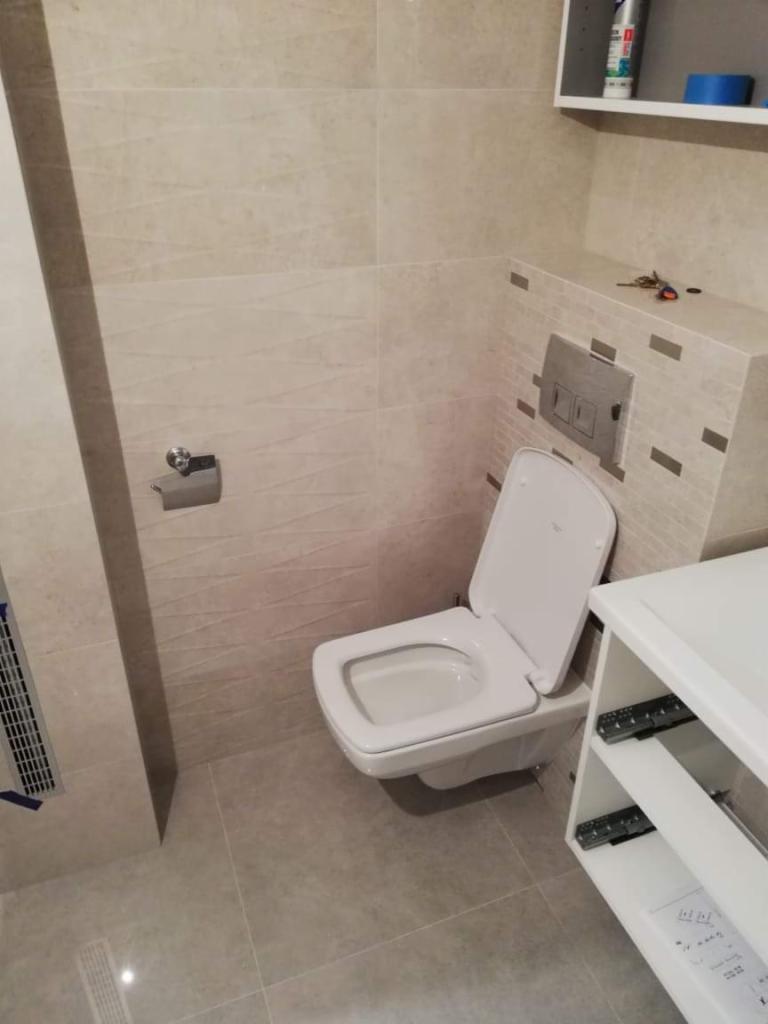 Nowoczesna toaleta podwieszana z otwartą klapą, beżowe płytki imitujące kamień na ścianach i podłodze, biała szafka wisząca z otwartymi półkami, klucze leżące na obudowie spłuczki.