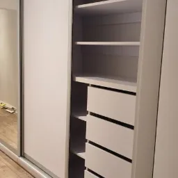 Biała szafa wnękowa z przesuwnymi drzwiami, widoczne półki i szuflady, drewniana podłoga.