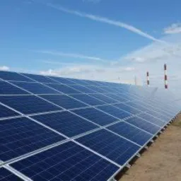 Rząd paneli słonecznych pod błękitnym niebem z białymi chmurami, w tle kominy z czerwono-białymi pasami i siatka ogrodzeniowa.