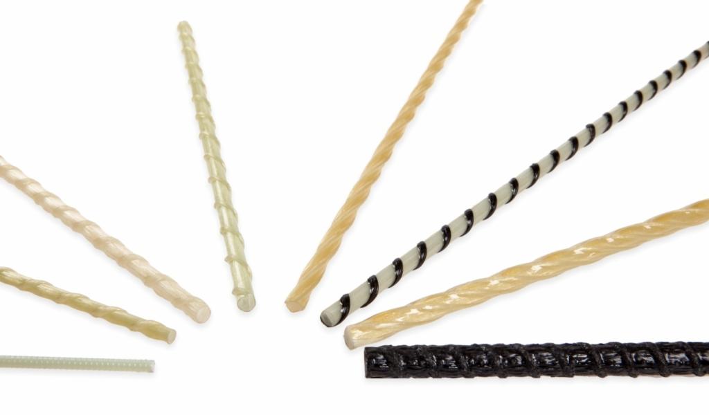 Kompozytowe pręty zbrojeniowe GFRP i BFRP o różnej kolorystyce i fakturze na białym tle, widoczne skręcone włókna i żebrowanie.