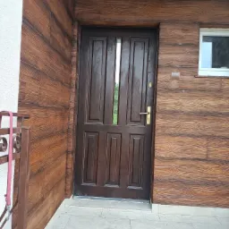 Elewacja budynku z brązową, drewnopodobną okładziną, ciemnobrązowe drzwi wejściowe z szybką, wisząca lampa zewnętrzna, okno z białymi ramami.