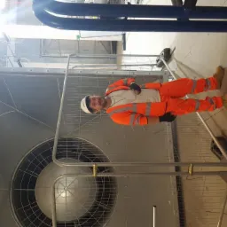 Pracownik w pomarańczowym kombinezonie i białym kasku stoi obok dużej, metalowej kratki wentylacyjnej w hali przemysłowej, zabezpieczonej barierkami.