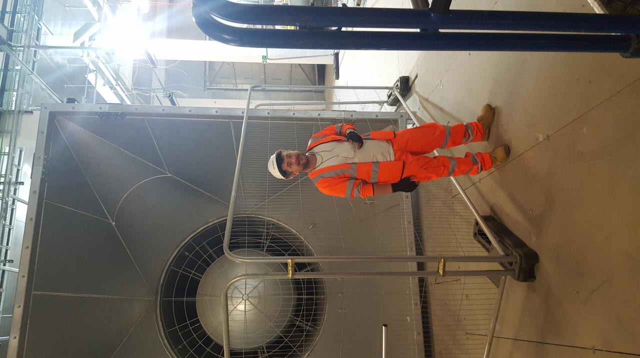 Pracownik w pomarańczowym kombinezonie i białym kasku stoi obok dużej, metalowej kratki wentylacyjnej w hali przemysłowej, zabezpieczonej barierkami.