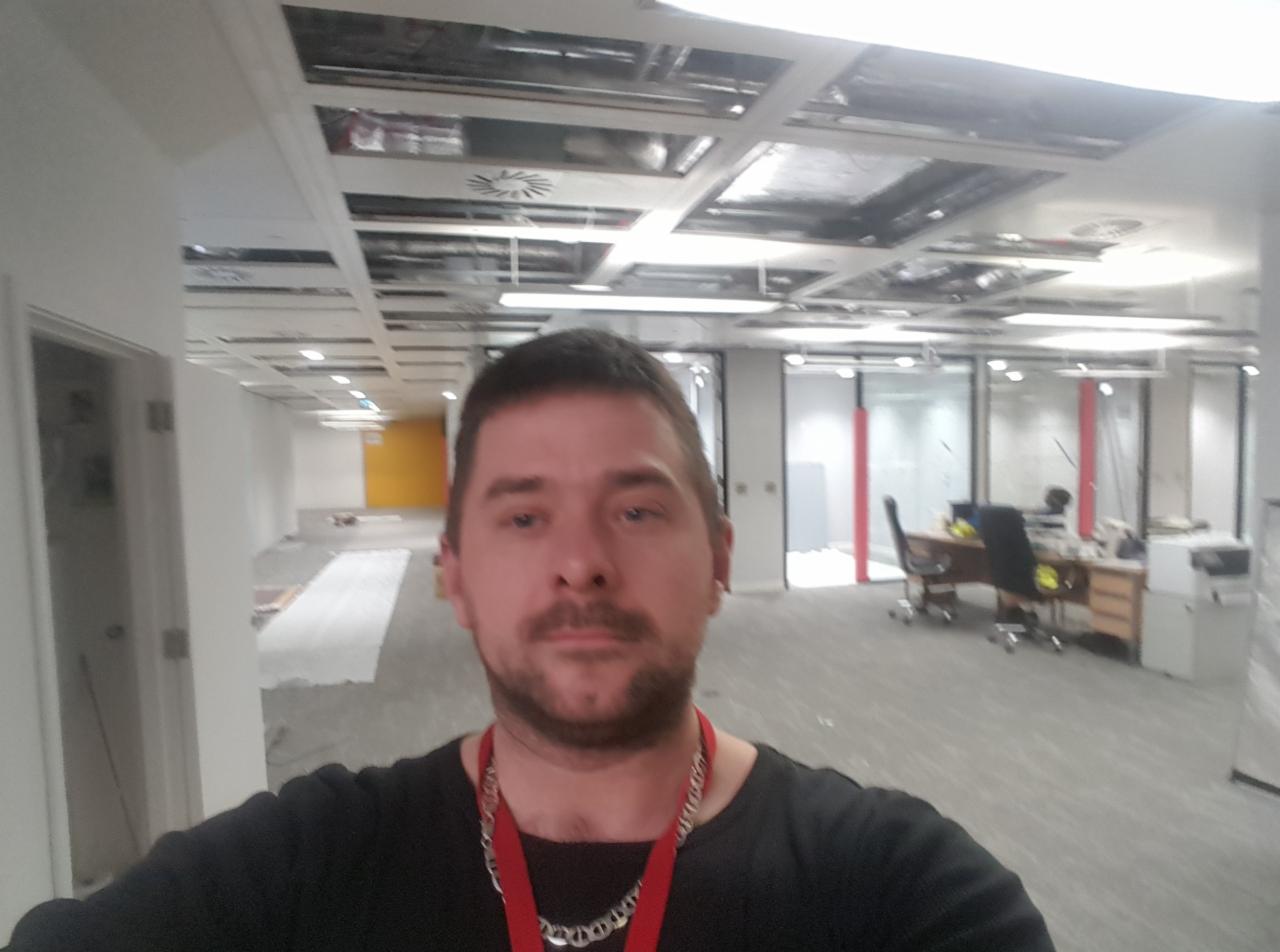 Mężczyzna robiący selfie na tle biura w trakcie remontu, widoczne niedokończone sufity podwieszane, puste przestrzenie biurowe i elementy instalacji elektrycznych.