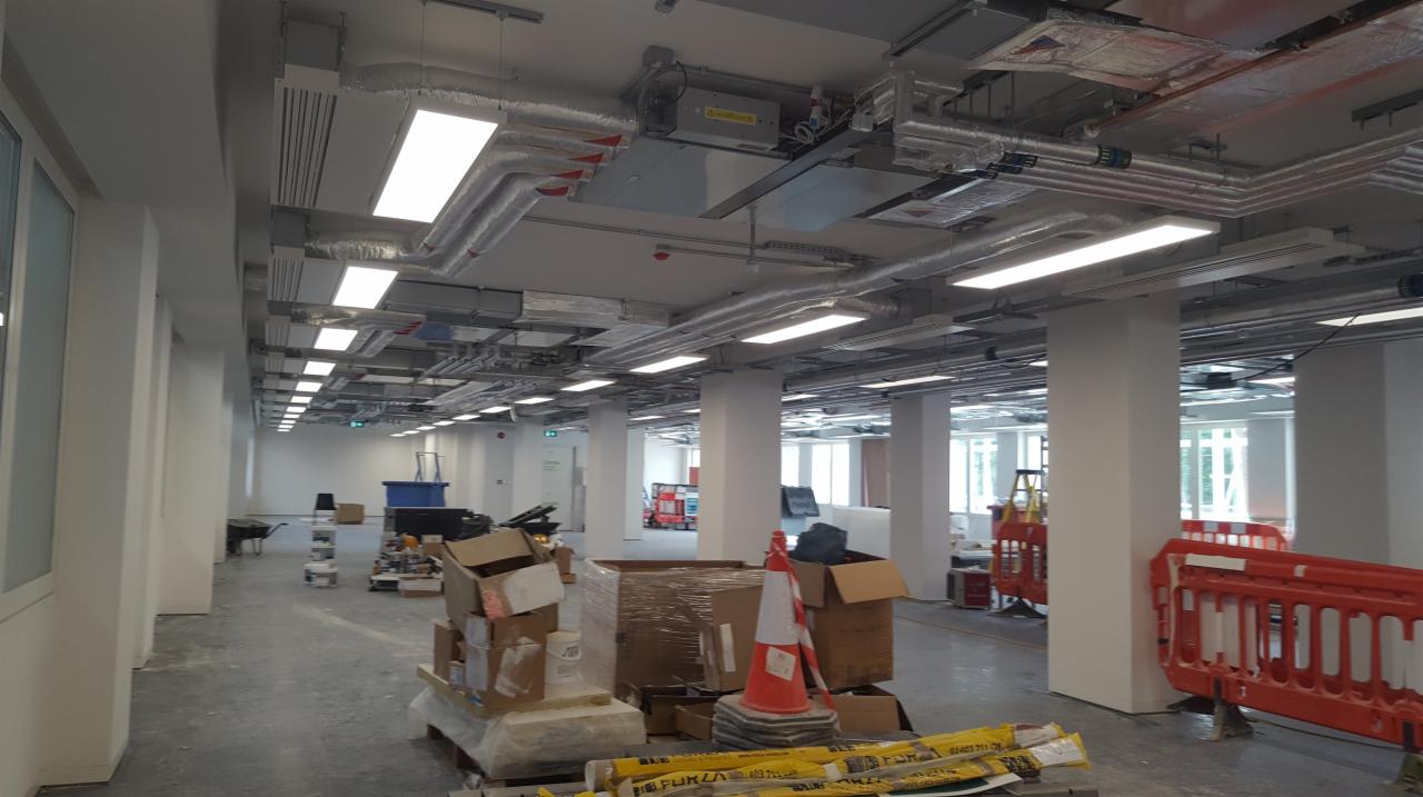 Montaż instalacji elektrycznej i wentylacyjnej w nowym biurze: widoczne kable, rury wentylacyjne, lampy sufitowe, kolumny nośne i materiały budowlane na podłodze.