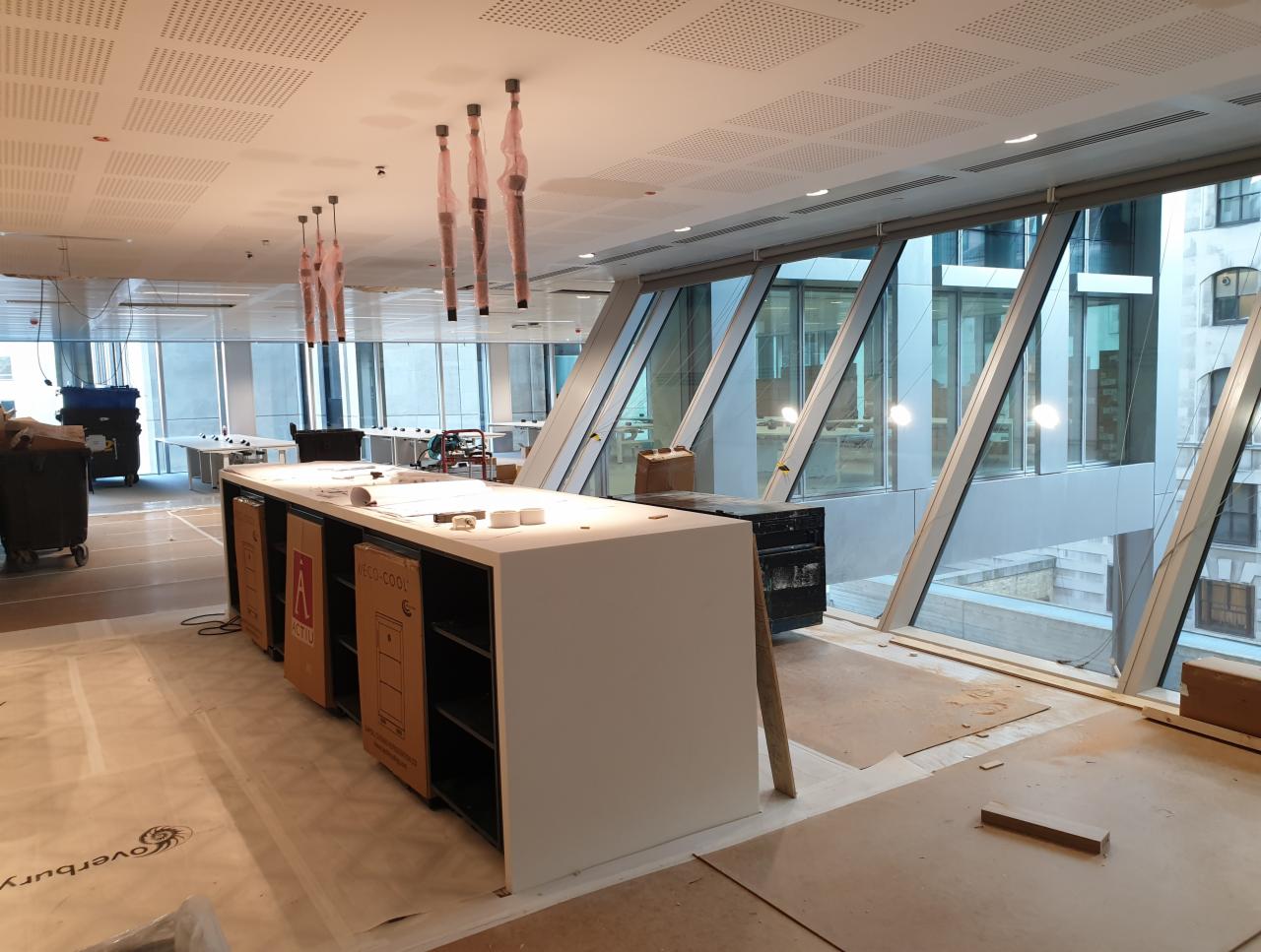 Wnętrze biurowe w trakcie prac instalacyjnych, widoczne kable zwisające z sufitu, zabudowa meblowa z kartonami urządzeń chłodniczych, duże okna z widokiem na budynki miejskie.