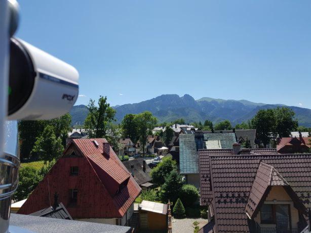 Kamera monitoringu IP marki POV zamontowana na zewnątrz budynku, z widokiem na panoramę miasta i góry w tle.