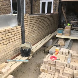 Robert Jasicki - Wznoszenie ściany z jasnej cegły na zewnątrz budynku, widoczne narzędzia murarskie, wiadro z zaprawą, poziomica oraz stos cegieł i karton soku na placu budowy.