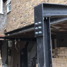 Robert Jasicki - Montaż stalowej konstrukcji wsporczej w otworze w ścianie z cegły, widoczne śruby mocujące i drewniane stemple podtrzymujące strop, cegły na palecie i drabina w tle.