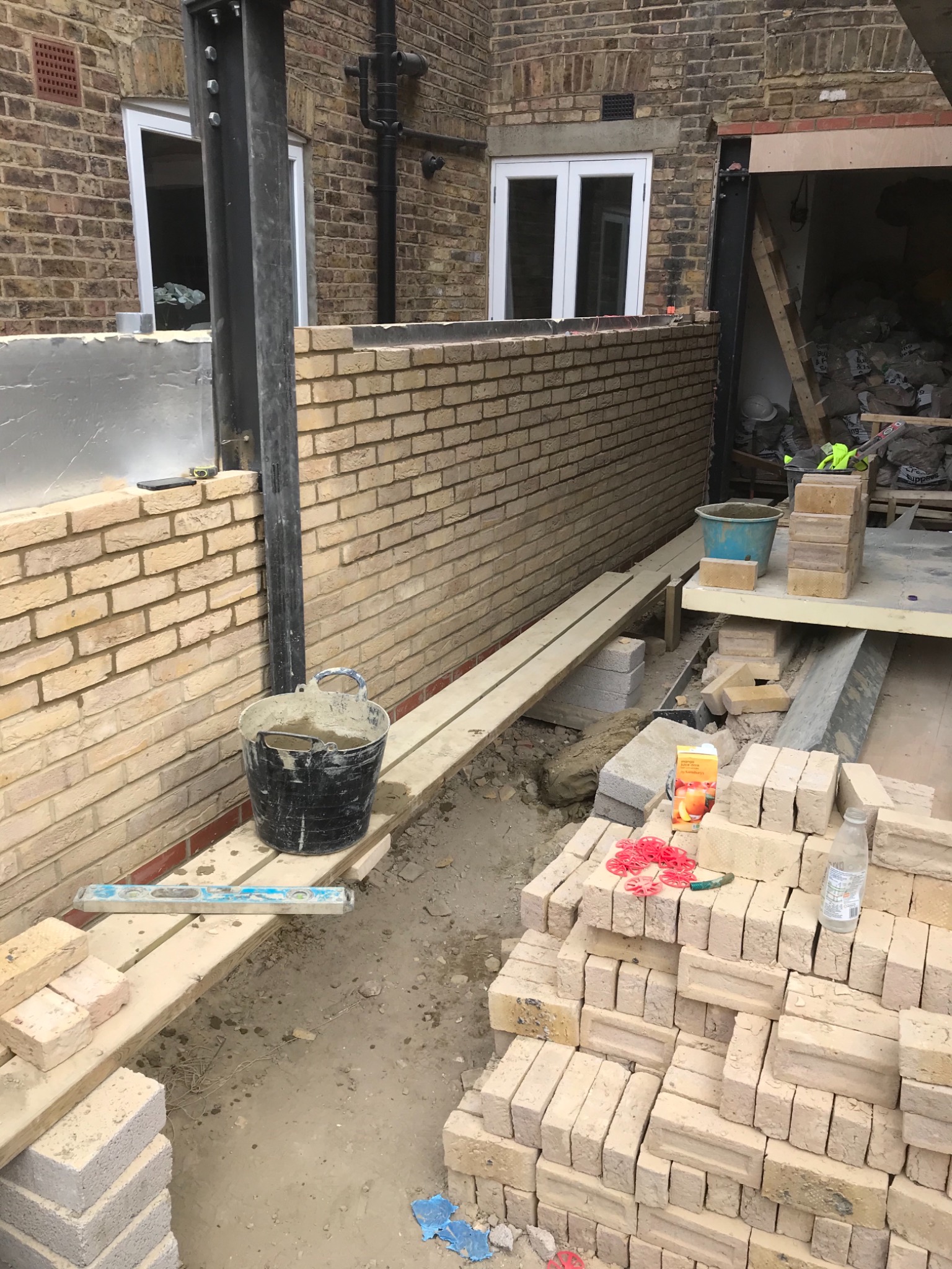 Wznoszenie ściany z jasnej cegły na zewnątrz budynku, widoczne narzędzia murarskie, wiadro z zaprawą, poziomica oraz stos cegieł i karton soku na placu budowy.