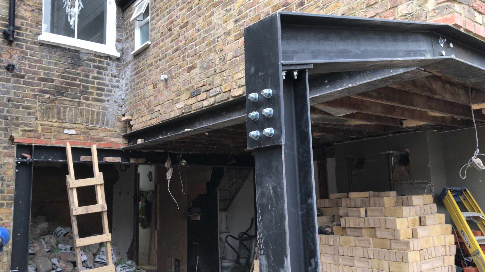 Montaż stalowej konstrukcji wsporczej w otworze w ścianie z cegły, widoczne śruby mocujące i drewniane stemple podtrzymujące strop, cegły na palecie i drabina w tle.