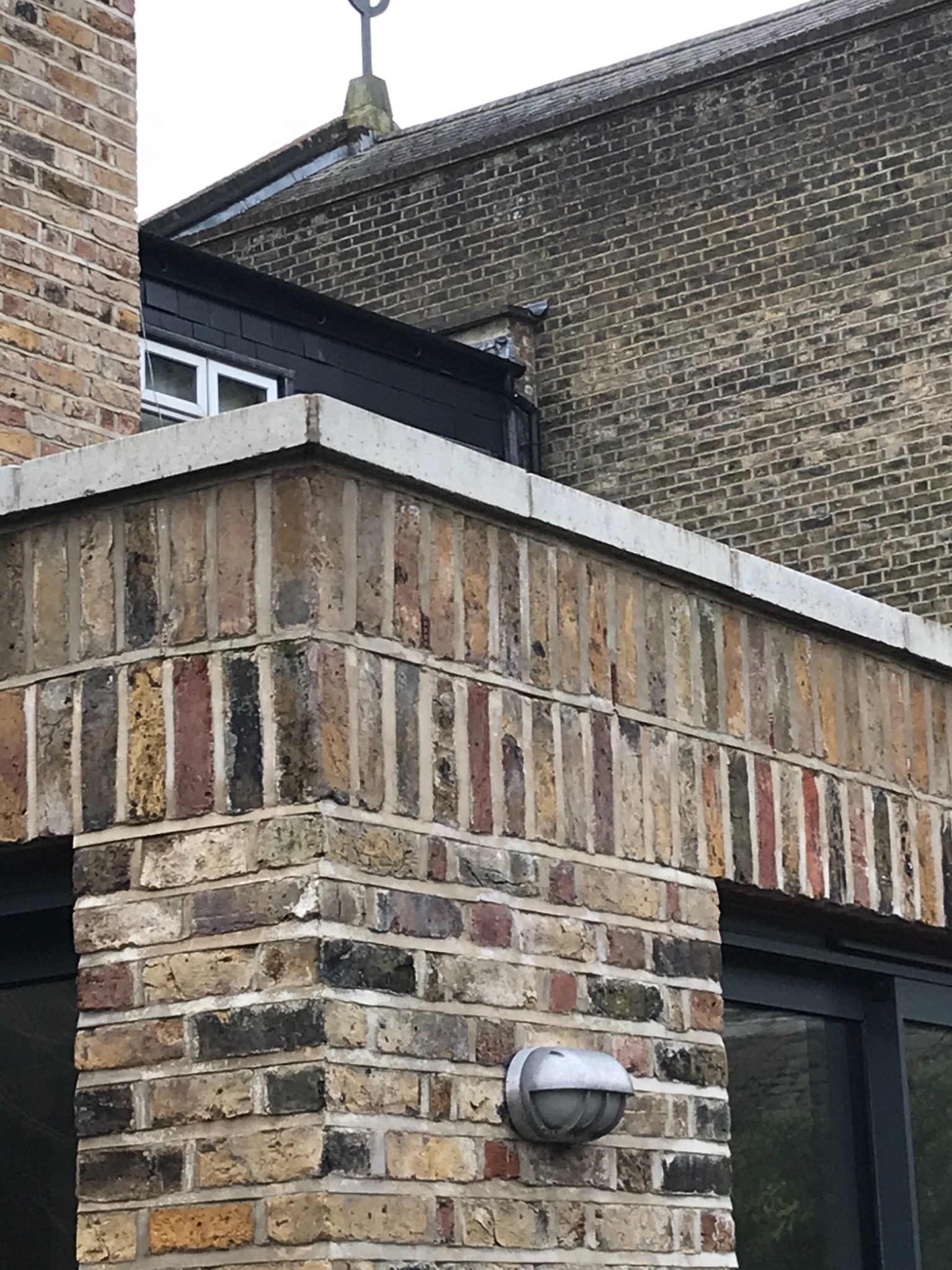 Fragment elewacji budynku z cegły klinkierowej w różnych odcieniach, z widocznym narożnikiem, oświetleniem zewnętrznym i fragmentem okna. Detal architektoniczny z elementami dekoracyjnymi.