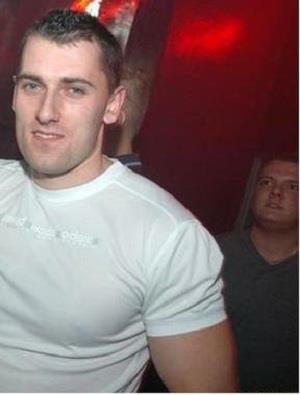 Mężczyzna w białej koszulce z krótkim rękawem na czerwonym tle, z innym mężczyzną w tle.