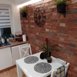 Jadalnia z ceglaną ścianą, zegarem, roślinami w czarnych doniczkach i białym stołem z dwoma miejscami nakrycia. Na stole dodatkowo roślina w doniczce i gazety. Krzesło przykryte niebiesko-żółtą...