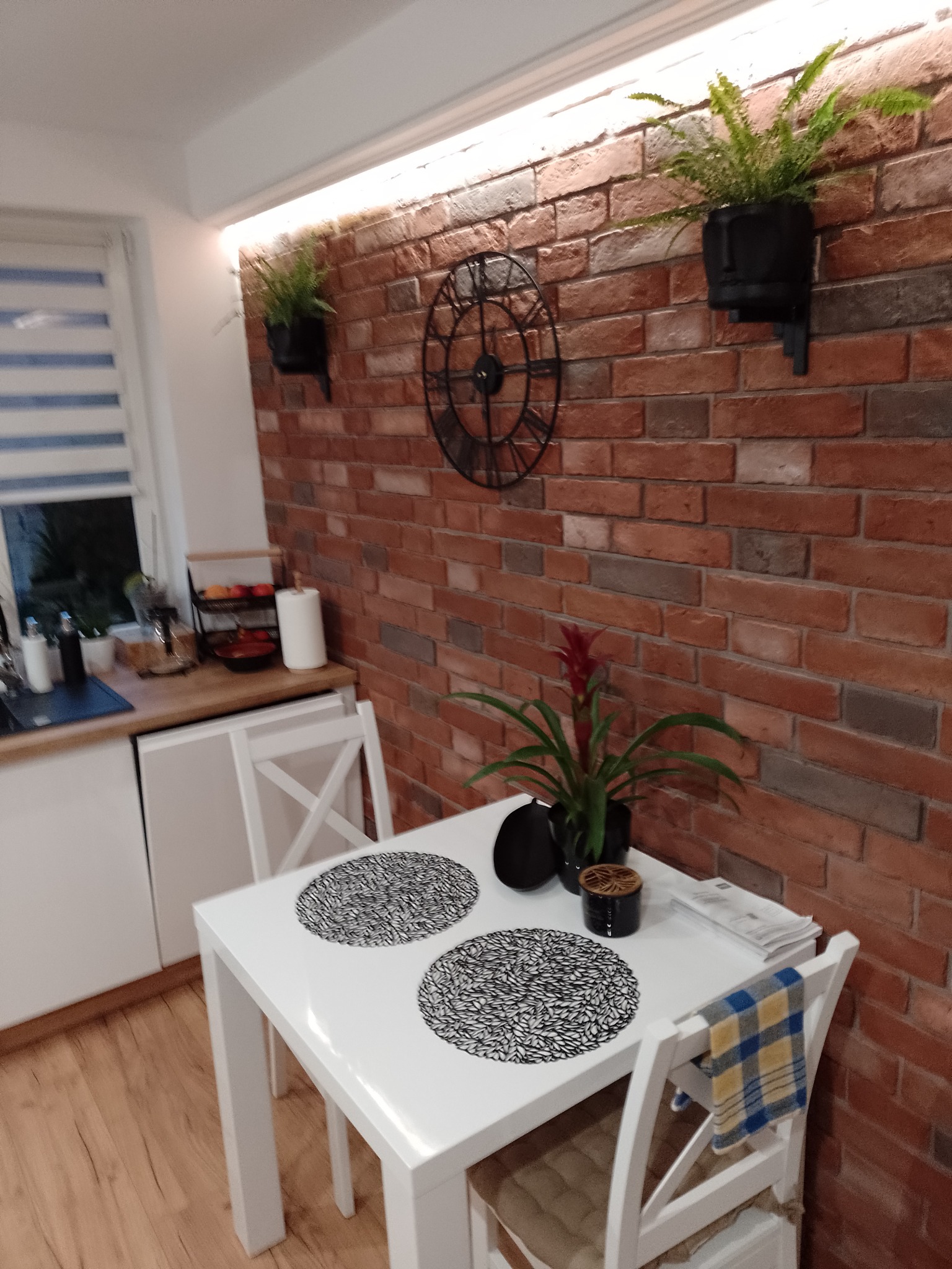 Jadalnia z ceglaną ścianą, zegarem, roślinami w czarnych doniczkach i białym stołem z dwoma miejscami nakrycia. Na stole dodatkowo roślina w doniczce i gazety. Krzesło przykryte niebiesko-żółtą...