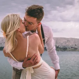 Para całująca się na tle morza i skalistych g&oacute;r; mężczyzna w okularach i szelkach trzyma kobietę w białej sukience z odkrytymi plecami.