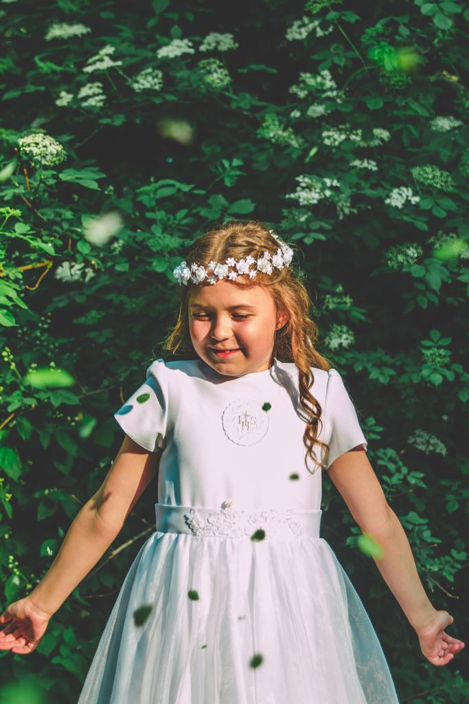 Dziewczynka w białej sukience komunijnej i wianku z białych kwiatów na tle zieleni, z opadającymi płatkami kwiatów.