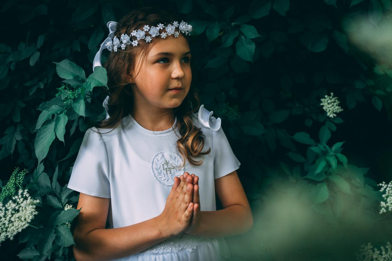 Dziewczynka w białej sukience i wianku na tle zieleni, złożone dłonie, uroczysty portret.