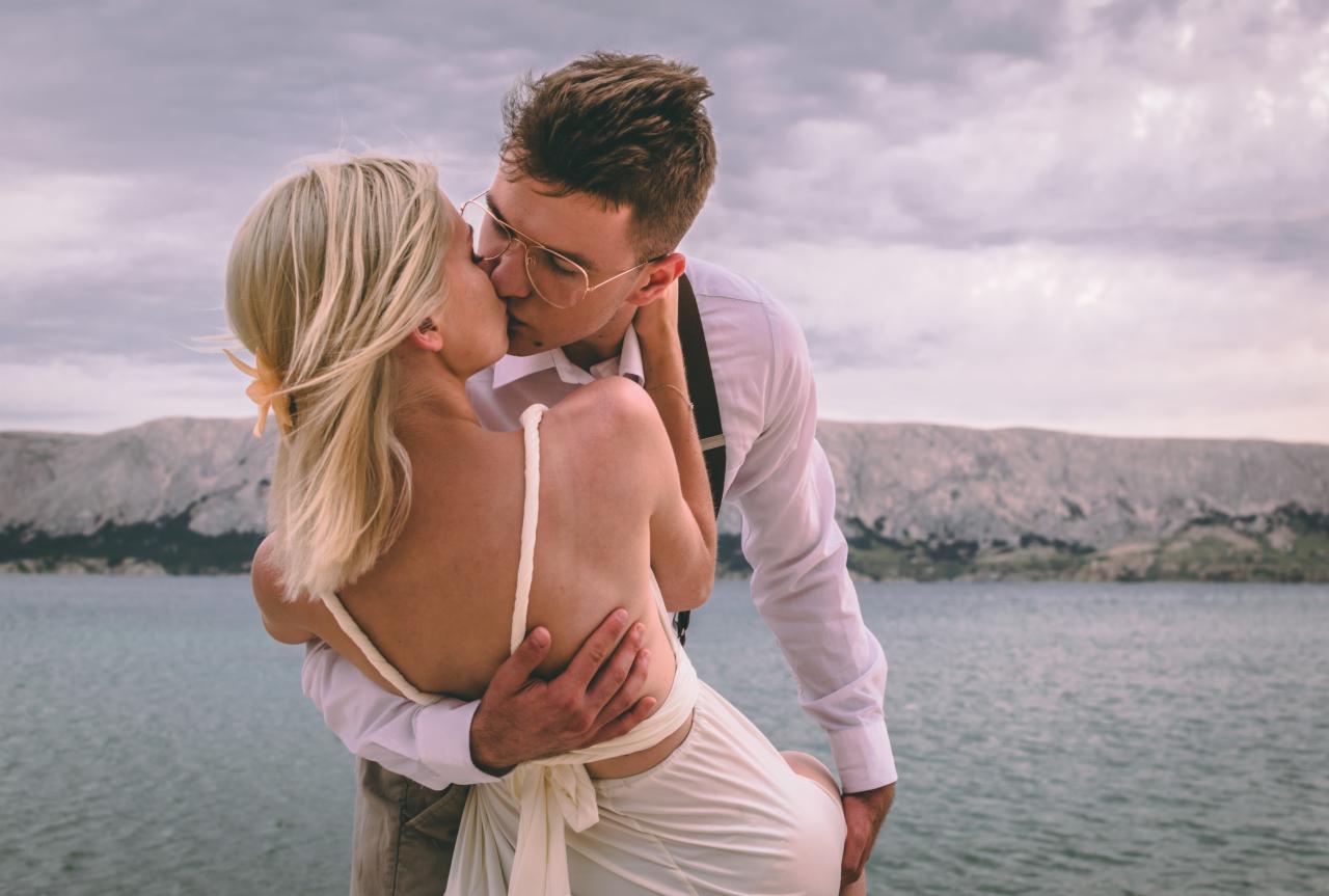 Para całująca się na tle morza i skalistych gór; mężczyzna w okularach i szelkach trzyma kobietę w białej sukience z odkrytymi plecami.