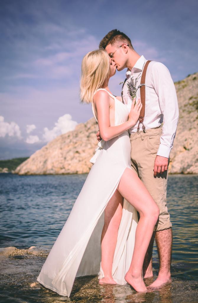 Para całująca się na tle morza i skalistego krajobrazu, kobieta w białej sukni z rozcięciem, mężczyzna w szelkach i krótkich spodniach, oboje boso w płytkiej wodzie.