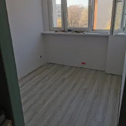 Pokój po położeniu paneli podłogowych w odcieniach szarości, widoczne białe ściany z otworami na gniazdka, okno z narzędziami na parapecie i widokiem na budynki