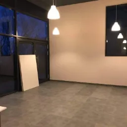 Szare płytki podłogowe ułożone w nowym pomieszczeniu z oknami i wiszącymi lampami, widoczna płyta gipsowo-kartonowa oparta o ścianę.