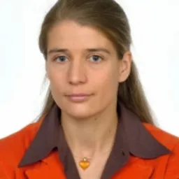 Portret kobiety w pomarańczowej marynarce i brązowej bluzce z wisiorkiem w kształcie serca na jasnym tle.