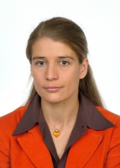 Portret kobiety w pomarańczowej marynarce i brązowej bluzce z wisiorkiem w kształcie serca na jasnym tle.