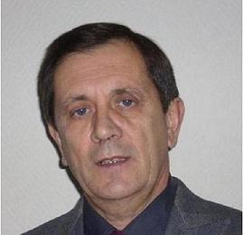 Portret mężczyzny w średnim wieku, z krótkimi, ciemnymi włosami, ubrany w ciemną koszulę i szarą marynarkę, na jasnym tle.