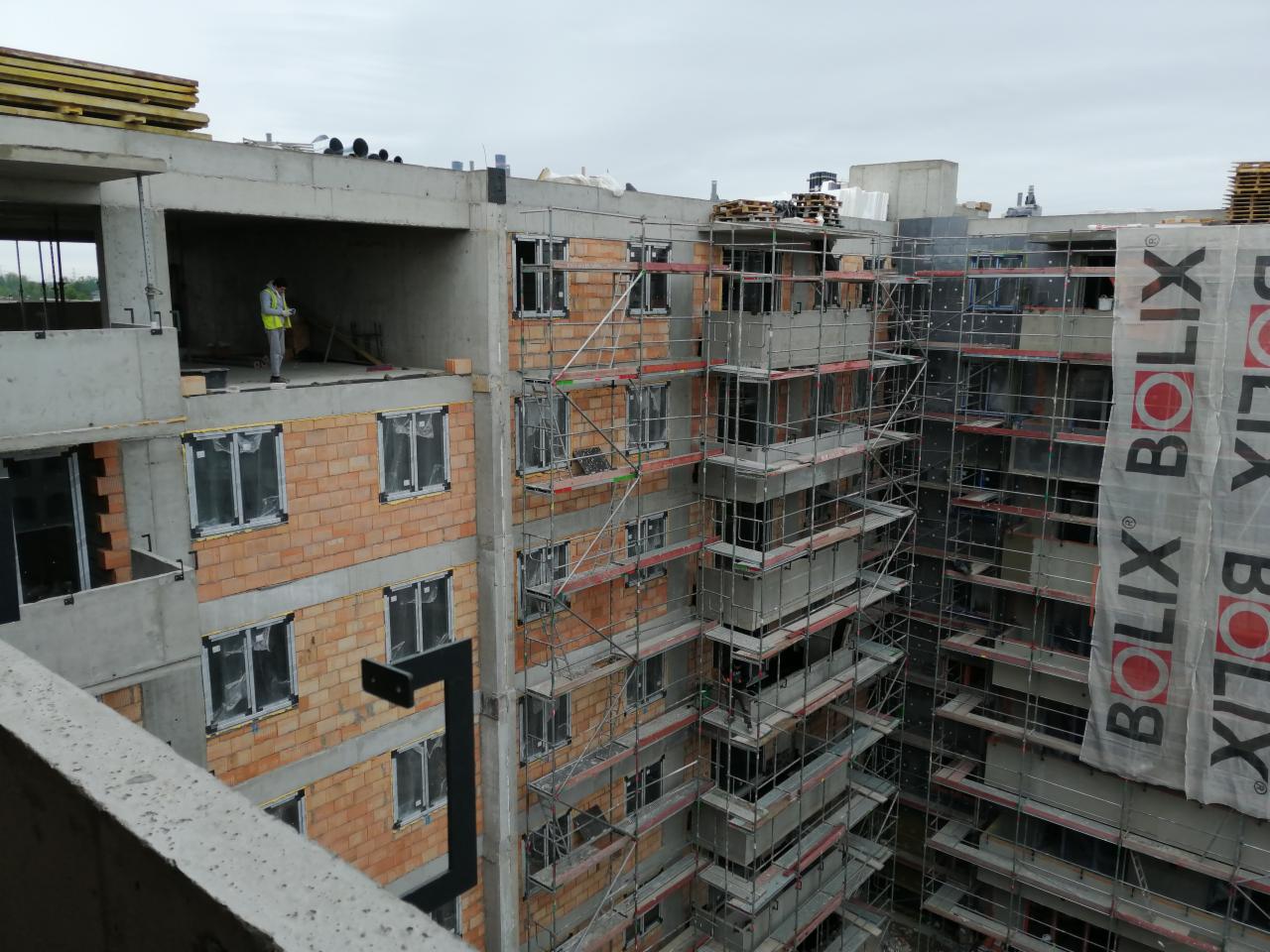 Budowa wielopiętrowego budynku z cegły, widoczne rusztowania i pracownik w kamizelce odblaskowej na jednym z pięter, ściany częściowo ocieplone styropianem, logo BOLIX na siatce rusztowania.