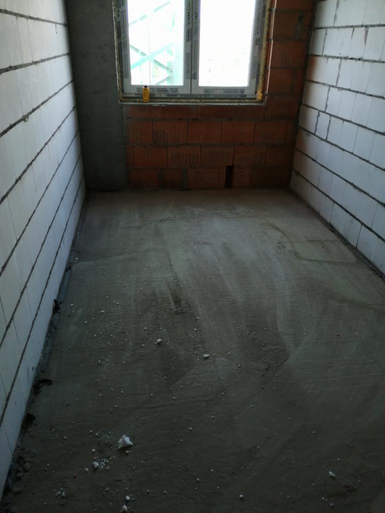 Surowy stan wnętrza: ściany z białych bloczków, fragment ściany z czerwonej cegły pod oknem, wylewka podłogowa, widoczne niedoskonałości i ślady prac budowlanych.