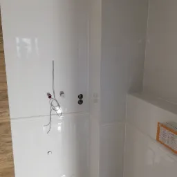 Biała, błyszcząca łazienka w trakcie remontu, widoczne wyprowadzone przewody instalacji wodnej i elektrycznej, częściowo ułożone płytki z dekoracyjnym frezem.