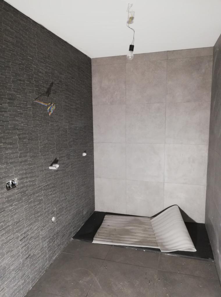 Łazienka w trakcie wykańczania: szare płytki ułożone na ścianach, widoczne wystające przewody elektryczne, podłoga przygotowana pod prysznic z zabezpieczającą folią w rogu.