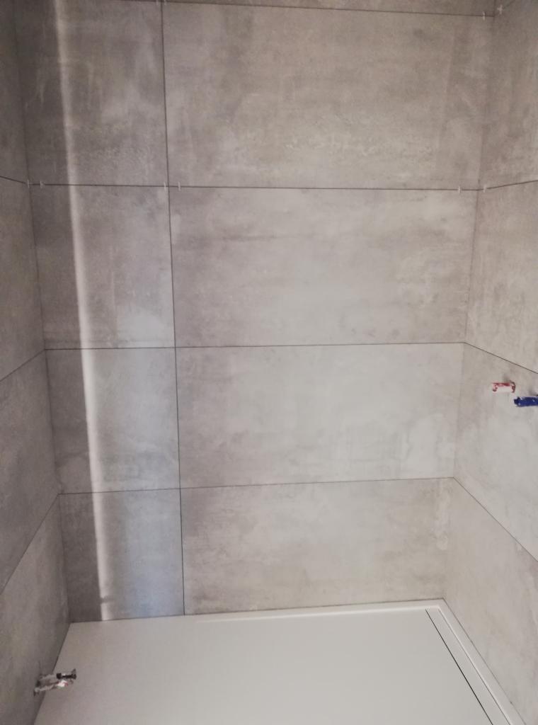 Ściana i narożnik wyłożone szarymi, matowymi płytkami ceramicznymi o dużym formacie, z widocznymi krzyżykami dystansowymi w spoinach, oraz fragment białe ościeżnicy drzwiowej w dolnej części kadru...