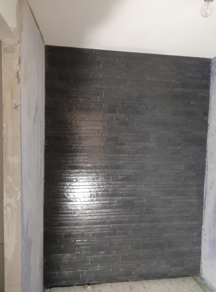 Ściana wykończona czarną, poziomą okładziną przypominającą łupek, z widocznymi refleksami światła, sąsiadująca z surową, nie wykończoną ścianą z widocznymi śladami zaprawy.