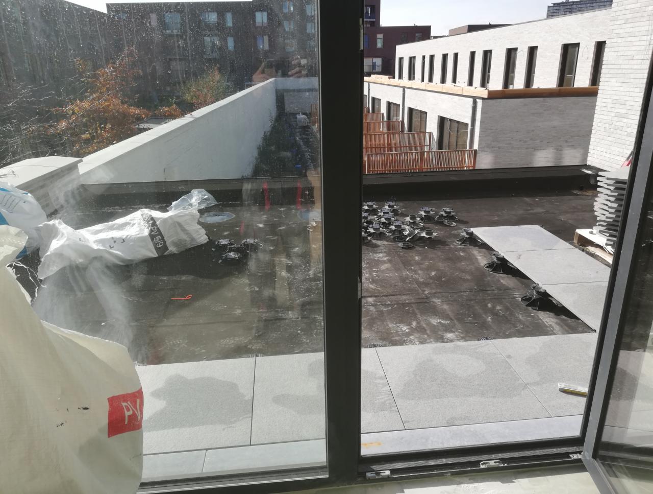 Montaż płytek na dachu: widok przez okno na częściowo wykończony dach z płytkami ułożonymi na regulowanych wspornikach, worki z materiałami budowlanymi w tle.