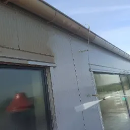 Czyszczenie elewacji budynku przemysłowego z widocznym brudem, strumień wody pod ciśnieniem z lancy, okna z odbiciem czerwonego przedmiotu, metalowe rynny.