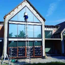 Pracownik myje okno w szczycie domu jednorodzinnego z użyciem wysokiej drabiny, dolne okna zabezpieczone folią ochronną.