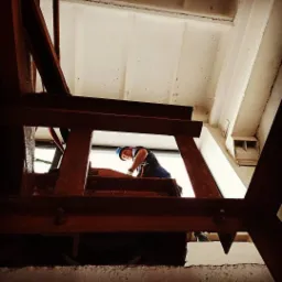 Pracownik w niebieskim kasku, zabezpieczony linami, wykonuje prace na wysokości, widoczny przez konstrukcję zardzewiałej metalowej ramy zębatki, z widokiem na okno i sufit.