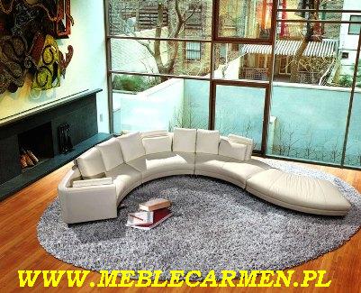 Panoramiczny, modułowy narożnik w odcieniach bieli, ustawiony na okrągłym, szarym dywanie shaggy, z książkami obok, na tle dużego okna z widokiem na zieleń.