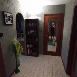 Korytarz z dekoracyjną szarą ścianą o nieregularnej fakturze, widoczny regał z książkami, lustro w drewnianej ramie odbijające osobę robiącą zdjęcie, oraz zielony wazon z kompozycją kwiatową.