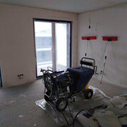 Dydziak sebastian firma remontowo-wykonczeniowa - Przygotowanie do szpachlowania maszynowego: agregat szpachlarski na folii, worki z gładzią oraz długie szpachelki przy ścianie w surowym wnętrzu z zabezpieczonymi oknami.