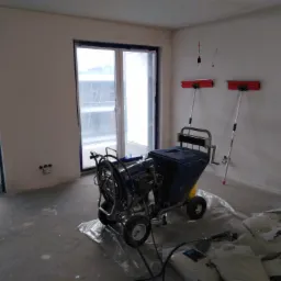 Przygotowanie do szpachlowania maszynowego: agregat szpachlarski na folii, worki z gładzią oraz długie szpachelki przy ścianie w surowym wnętrzu z zabezpieczonymi oknami.